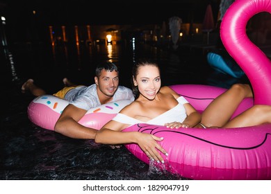 Selective Focus Of Young Couple Looking At Camera While Swimming On Rings In Pool At Night