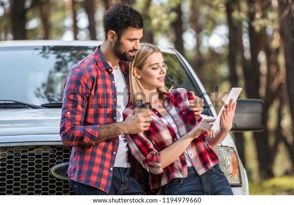 selective focus of young man with paper coffee cup\
standing near girlfriend while she using digital tablet near pick\
up car outdoors 