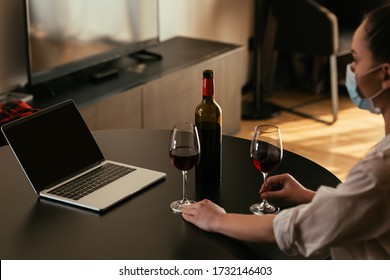 Selective Focus Of Young Woman In Medical Mask Near Laptop With Blank Screen, Glasses And Bottle Of Red Wine