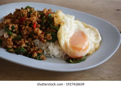 Selective focused, fried egg on top of fried basil leave with pork on steamed rice in plate, placed on wooden table. Thai popular food.  Thai food, popular food, fried egg, fast food concept.