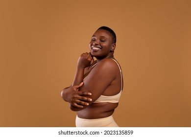 Self Acceptance, Love And Care. Portrair of half-turned smiling curvy African American woman with perfect skin and closed eyes embracing herself isolated on brown studio background, free copy space