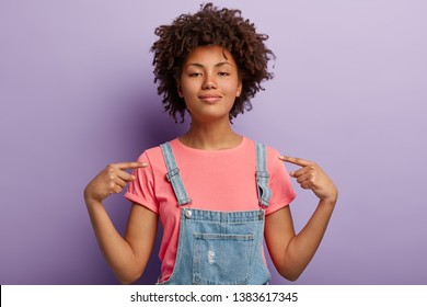 Self assured beautiful African American woman feels proud of her deeds, points at herself feels surge of pride, raises head, has dark healthy skin, wears casual clothes, isolated over purple wall