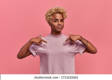 Self assured proud African American woman has blonde hairstyle satisfied with her own high achievements, points with two hands at herself, or at a t-shirt for printing, isolated over pink background.