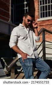 Self Assured Stylish Young Male In Smart Casual Clothes Holding Hand In Pocket And Adjusting Sunglasses, While Leaning On Banister On Sunny Day Outside Brick Building