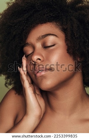 Self assured young ethnic female millennial with curly Afro hair and perfect skin touching face with closed eyes, during beauty routine