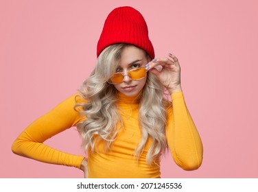 Self assured young female millennial with long blond hair in casual clothes, and knitted hat adjusting trendy sunglasses and looking at camera against pink background - Shutterstock ID 2071245536