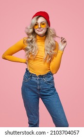Self assured young female model in trendy sunglasses and hat, touching long blond hair and smiling while standing against pink background with hand on waist - Shutterstock ID 2103704057