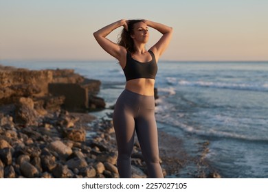 Self assured young fit woman in activewear touching long wavy hair, while standing in rocky beach near ocean with closed eyes after practicing yoga at sunset - Shutterstock ID 2257027105