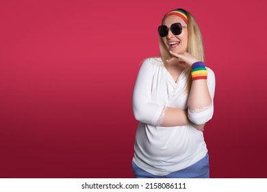 Self Assured Young Homosexual Female In Sunglasses And Colorful Pride Wristband And Headband Showing Tongue And Touching Chin Against Pink Background