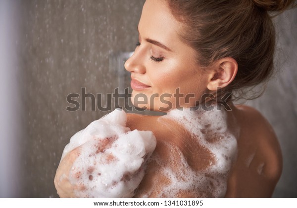 Self care treatment concept. Close up back side\
portrait of pretty smiling woman closing eyes while enjoying shower\
with foam at home