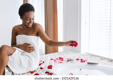 Self care treatment, time for herself, enjoying peace and calm. Cheerful millennial african american lady sits on bath, throws flower petals, in bathroom with candles interior in morning, copy space