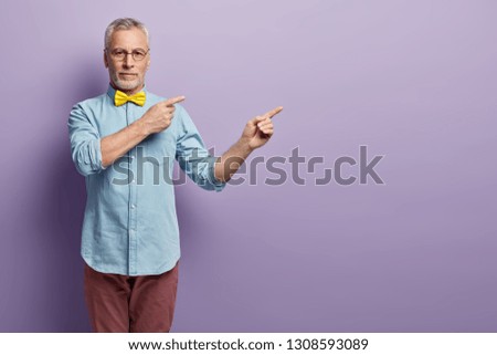 Self confident assertive grandfather points right, wears yellow bowtie, stylish denim shirt and trousers, poses over purple background with empty space for your advertsing content or promotion