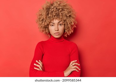 Self confident curly haired woman raises eyebrows has serious expression poses with arms folded against red background listens attentively interlocutor. Beautiful female model in assertive pose