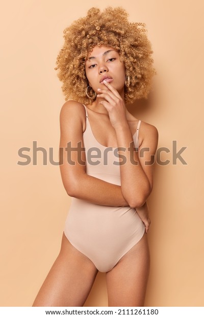Self confident curly haired young woman looks\
seriously at camera touches face looks directly at camera wears\
bodysuit has good figure isolated over beige background. Women and\
body care concept