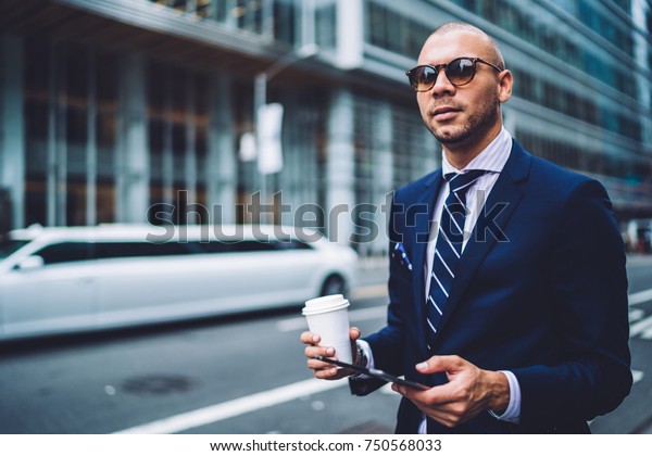Self confident executive manager in trendy\
sunglasses standing on road with coffee takeaway waiting for cab,\
handsome businessman in formal wear using application on touchpad\
for calling taxi service\
