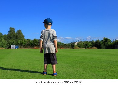 Self Confident Kid Standing Up At A Green Lawn. Gray Shirt And Shorts With A Blue Cap. Back Towards Camera. Nice Summer Day Outside. Stockholm, Sweden, Europe.