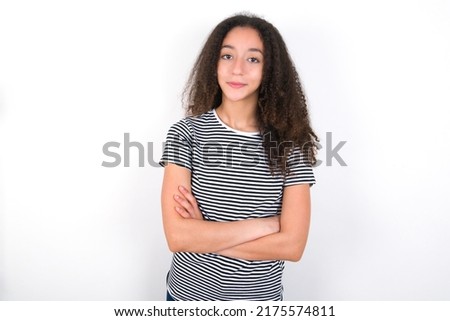 Self confident serious calm young beautiful brunette woman wearing striped t-shirt over white wall stands with arms folded. Shows professional vibe stands in assertive pose.