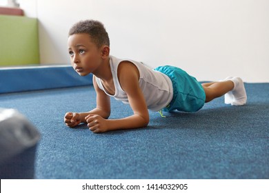 Self Determined African American Boy Dressed In Sports Clothes Planking At Gym, Feeling Tired. Indoor Image Of Black Male Kid Doing Elbow Plank On Floor At Gym. Endurance And Strength Concept