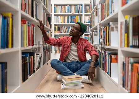 Self education. Young African American guy sits on floor in library between bookshelves, spending time at bookstore, searching for book, black male student preparing for college test. World book day