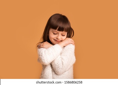 Self Love, Little Child Pretty Girl Hugging Herself. Comfort Zone, Pleasure, Cozy Concept. Cheerful Charming Caucasian Child In White Sweater Over Orange Background.