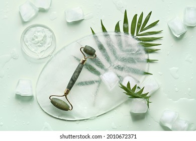 Self made moisturizer and green jade face roller on oval piece of ice and ice cubes. Exotic palm leaves on mint green background. Minimal flat lay, top view. Facial massage, handmade cosmetics.