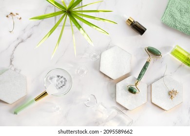 Self made moisturizer and green jade face roller on marble hexagons. Exotic sedge water plant leaf on off white marble backdrop. Facial massage, handmade cosmetics concept backdrop.