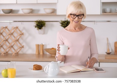 Self Organisation. Cheerful Senior Smiling Woman Making Notes And Standing In The Kitchen While Drinking Tea