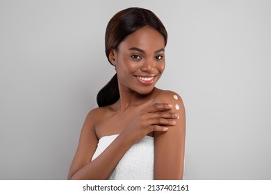 Self Pampering Concept. Beautiful Black Lady Applying Body Cream On Shoulder, Happy African American Woman Wrapped In Towel Using Moisturising Skin Lotion After Bath, Gray Background, Copy Space - Powered by Shutterstock