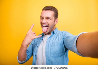 Self Portrait Of  Attractive, Crazy Man With Bristle, Stubble In Jeans Shirt, Shooting Selfie And Showing Rock And Roll Symbol With Tongue Out To The Front Camera Over Yellow Background