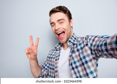 Self portrait of bearded, cool, mad, brunet guy in casual outfit, checkered shirt showing v-sign to the camera, laughing, shouting, screaming over grey background