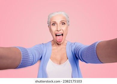 Self portrait of gray haired beautiful smiling funky funny playful old lady wearing casual, grimacing, showing tongue out, isolated over violet background