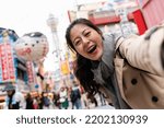 self portrait of happy Asian Japanese female tourist looking at camera while taking picture with tsutenkaku tower at background in nostalgic Shinsekai shopping area of Osaka Japan