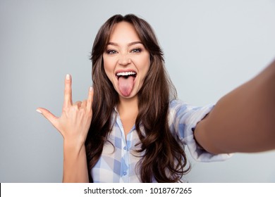 Self Portrait Of Nice, Attractive, Crazy Woman, Shooting Selfie And Showing Rock And Roll Symbol With Tongue Out To The Front Camera Over Grey Background