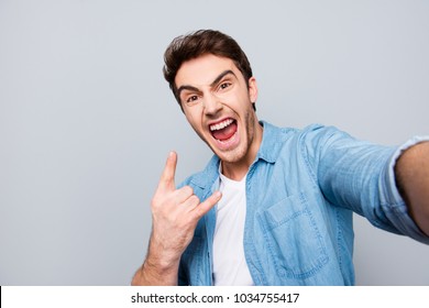 Self Portrait Of Shouting, Foolish, Individual, Attractive, Crazy, Funny Man With Wide Open Mouth, Stubble In Jeans Shirt Shooting Selfie On Front Camera, Showing Rock And Roll Sign On Grey Background