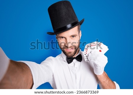 Self portrait of stylish cheerful joyful illusionist with stubble in tophat shooting selfie on front camera of smart phone, having set of cards in hands, isolated on grey background
