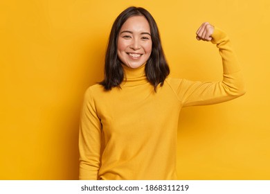 Self proud cheerful Asian woman raises arm and shows biceps has happy expression demonstrates her achievements after training in gym dressed casually isolated on yellow background. Look at my muscles