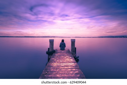 Self reflection in magical world of fantasy. One woman sits on a wooden pier. Cloudy above the lake. Long exposure
