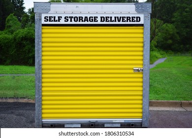 A Self Storage Pod Or Shed With A Yellow Door And Lock. Sign Indicates A Deliverable Self-storage Unit. Unit Is On A Street Or Parking Lot With Green Grass And Trees In Background. Outdoors In Daytime