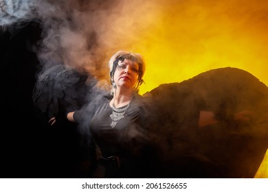 Self-assured Adult Blonde Middle Age Woman With Black Angel Wings. Model Posing In Studio On Black Background. Dangerous Cupid On Valentine's Day