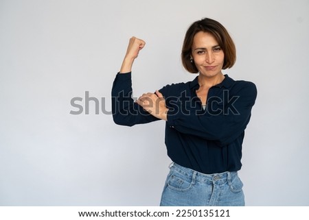 Self-assured Caucasian woman showing bicep. Portrait of proud mature female model with short dark hair in navy-blue shirt looking at camera, smirking, demonstrating power. Strength, success concept