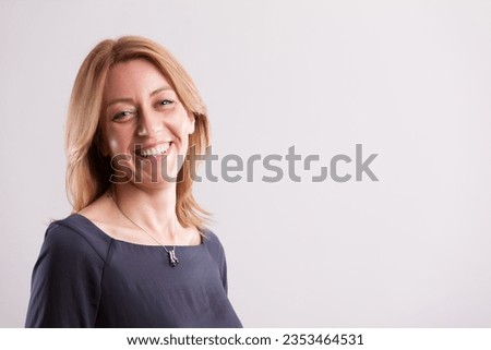 self-assured middle-aged blonde woman around 40, clad in a gray-blue dress, radiates happiness and contentment. Recognizing life's challenges and valuing self-reliance, her professional prowess shines