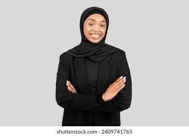 Self-assured millennial African American businesswoman with a bright smile, wearing a hijab and a black blazer, crosses her arms in a stance of confidence and professional poise - Powered by Shutterstock