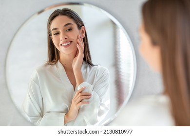 Self-Care Concept. Portrait Of Attractive Young Female Looking At Mirror, Beautiful Woman Wearing White Silk Robe Touching Soft Skin On Face And Smiling, Enjoying Her Reflection, Selective Focus - Shutterstock ID 2130810977