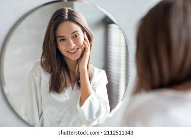 Self-Care Concept. Young Attractive Woman Looking At Mirror In Bathroom, Pretty Female Touching Face And Smiling To Reflection, Enjoying Her Beautiful Smooth Skin, Selective Focus, Closeup - Powered by Shutterstock