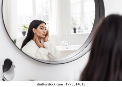 Selfcare Concept. Young Charming Asian Woman In White Bathrobe Touching Her Soft And Healthy Skin, Checking Wrinkles While Sitting In Front Of Big Mirror In Bathroom At Her Apartment