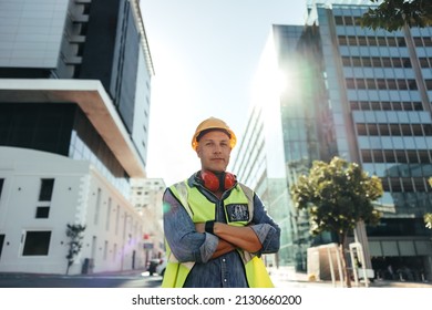 Self-confident Construction Worker Looking At The Camera While Standing With His Arms Crossed In The City. Mid-adult Blue Collar Worker Standing In Front Of High Rise Buildings In His Workwear.