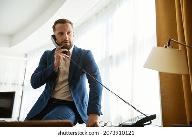 Self-confident Handsome Businessman, Attractive Man, Prosperous Financial Investor On A Business Trip, Sitting On The Office Table Corner In The Hotel Room And Calling The Front Desk By Landline Phone