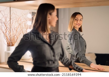 Self-confident Woman looking at her reflection into the mirror indoors. Beautiful interior design