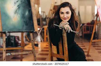 Self-employed young painter looking at the camera cheerfully while sitting on a chair in her workshop. Happy female artist holding a paintbrush with colour painted hands.