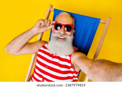 Selfie of aged smiling positive pensioner wear striped summer swimsuit touch sunglasses at beach sunbed isolated on yellow color background - Powered by Shutterstock